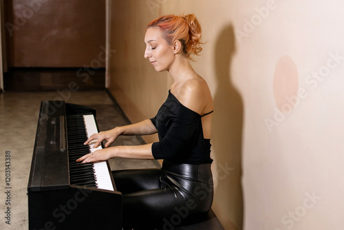Young girl woman in black clothes playing piano her favorite classical music, learning new melody, taking lessons. Romantic subdued light with shadow against beige wall. Side view. Copy space