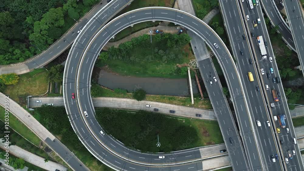 Drone footage of a busy highway interchange with circular ramps and heavy traffic flow. Lush greenery surrounds the roads.