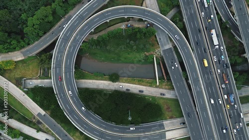 Drone footage of a busy highway interchange with circular ramps and heavy traffic flow. Lush greenery surrounds the roads.