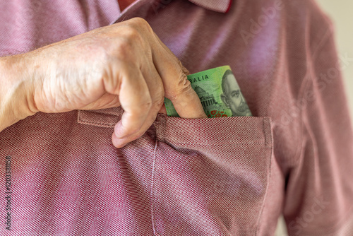 Close-up on a man's hand taking a one hundred Australian dollar note into his shirt's pocket. Concept illustration for bribing, corruption or cash payment.