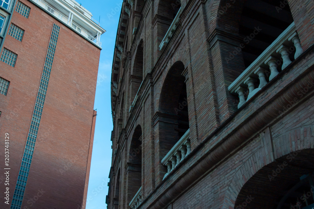 Fototapeta premium Brick facade of historic bullring las arenas contrasting with modern red building, glass windows reflecting blue sky in barcelona urban landscape