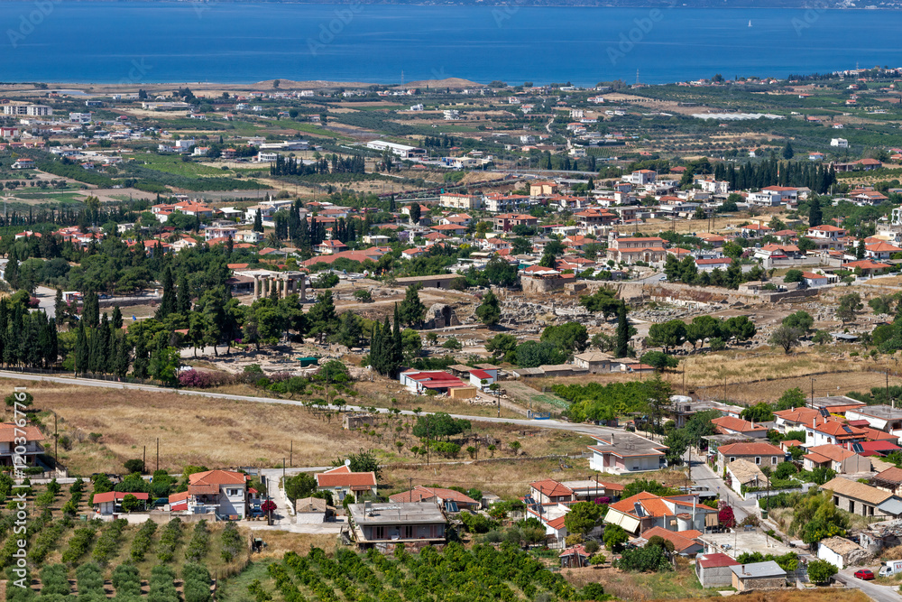 Aerial view of Ancient Corinth, Greece, showcasing the Temple of Apollo, historical ruins, traditional architecture, and the scenic Gulf of Corinth in the background.