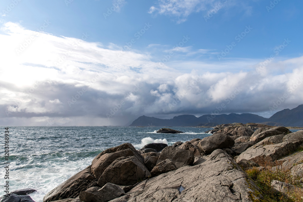 Low angle view of Southern coastline of the island of Austvagoya on Lofoten island chain archipelago in Norway with rain clouds on coastal mountain range and bright sun and blue sky Low angle view of Southern coastline of the island of Austvagoya on Lofoten island chain archipelago in Norway with rain clouds on coastal mountain range and bright sun and blue sky