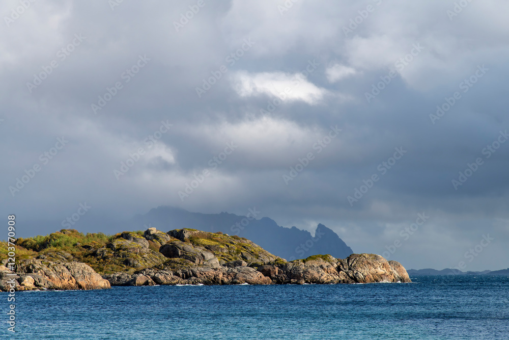 Dramatic cloudscape with dark cloud cover over silhouette mountain range and rocks in bright sun along fjord on island of Austvagoya on Lofoten island chain archipelago in Norway Dramatic cloudscape with dark cloud cover over silhouette mountain range and rocks in bright sun along fjord on island of Austvagoya on Lofoten island chain archipelago in Norway