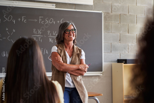Smiling female mature teacher standing in front of a blackboard in high school classroom, teaching chemistry while students listen attentively in a engaging atmosphere. image with copy space.