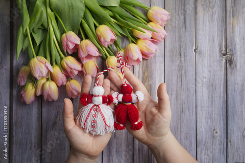Martenitsa, Martisor in female hands against the background of a bouquet of pink tulips on a wooden table top view