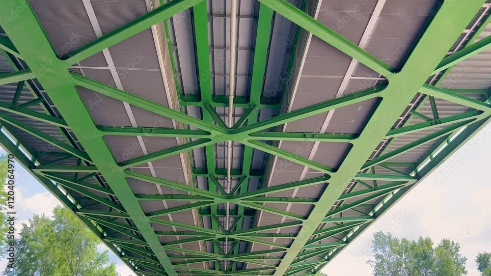 A close-up view of the underside of a green steel bridge, showcasing its geometric structure and engineering details. The scene emphasizes symmetry and industrial design.