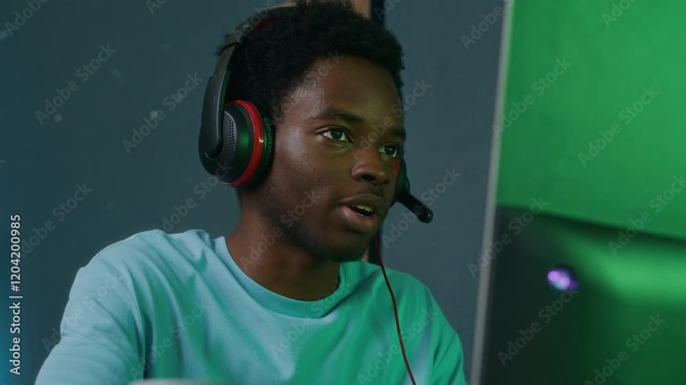 Medium close up shot of happy gen Z man of African American ethnicity wearing headset and speaking on mic while streaming during online game session on computer indoors