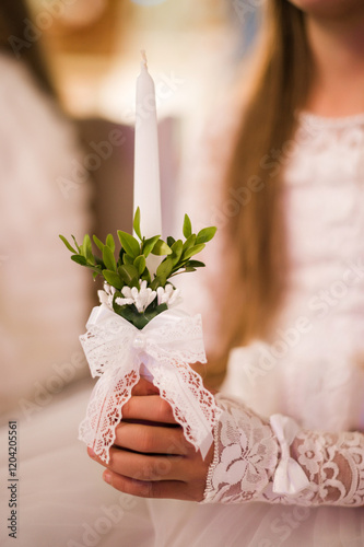 Close-up of a young girl's hand holding a white candle adorned with greenery and lace. The image evokes feelings of innocence, faith, and celebration. photo