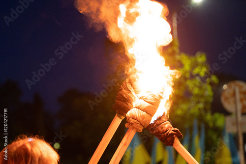 Two adults hold aloft flaming torches in the evening.  The light illuminates their faces and the background subtly. A sense of community and shared purpose is palpable.