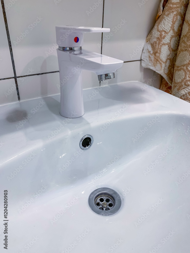 Fototapeta premium White faucet on a white sink in the bathroom, close view, white tile on the wall with black grout, towel in the distance. High quality photo