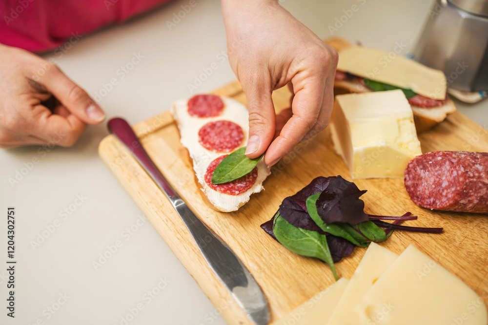 Woman preparing breakfast in a kitchen, making a sandwich with butter, sausage, cheese, and spinach leaves on bread, on a table Woman preparing breakfast in a kitchen, making a sandwich with butter, sausage, cheese, and spinach leaves on bread, on a table