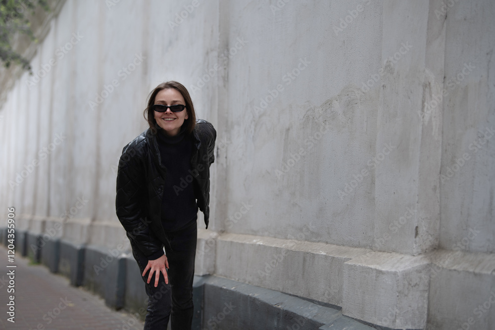 Obraz premium A woman smiles while wearing a black leather jacket and sunglasses, playfully bending next to a weathered white wall in an urban setting on a sunny day