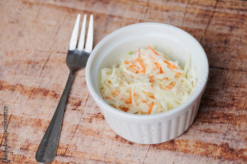 A small bowl of coleslaw sits on a wooden surface next to a fork ready for serving.
