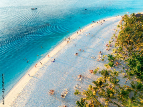 Aerial view of green palm trees, umbrellas on the sandy beach of Indian Ocean at sunset. Summer holiday in Kendwa, Zanzibar island. Tropical landscape with palms, white sand, clear blue sea. Top view photo