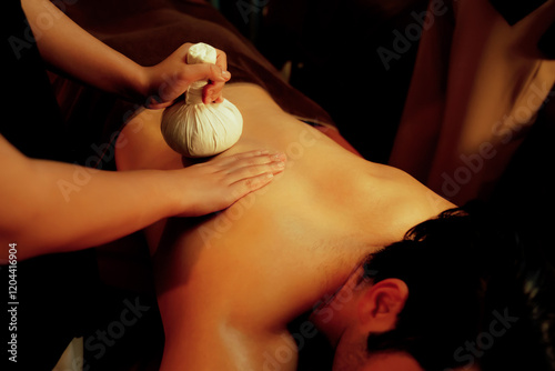 Hot herbal ball spa massage body treatment, masseur gently compresses herb bag on man body. Tranquil and serenity of aromatherapy recreation in warm lighting of candles at spa salon. Quiescent photo