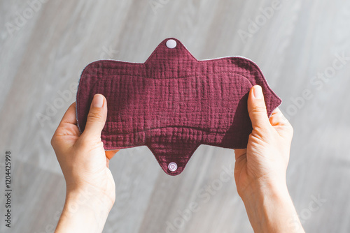 Reusable burgundy fabric muslin menstrual pad held in woman hands, sustainable and eco-friendly period product design, with neutral background, concept of ecology women's self-care during menstruation