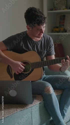Vertical video: Young man is sitting with a guitar, a concert for advertising. Guy is learning to play the guitar and watching a music lesson on his laptop
