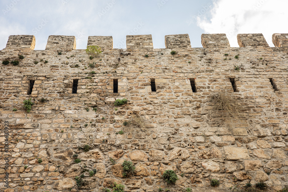 Ancient stone fortress wall with battlements and arrow slits under blue sky Ancient stone fortress wall with battlements and arrow slits under blue sky