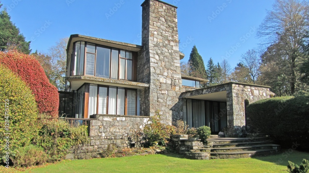Modern stone house with curved walls, large windows, and a chimney, situated in a landscaped garden. Modern stone house with curved walls, large windows, and a chimney, situated in a landscaped garden.