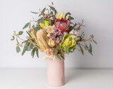 Beautiful floral arrangement of mostly Australian native flowers in a pink vase, including protea, banksia, eucalyptus leaves and wax flowers, on a white table with a white background.