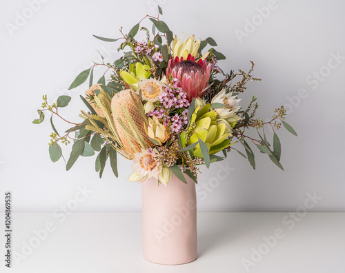Fototapeta Naklejka Na Ścianę i Meble -  Beautiful floral arrangement of mostly Australian native flowers in a pink vase, including protea, banksia, eucalyptus leaves and wax flowers, on a white table with a white background.