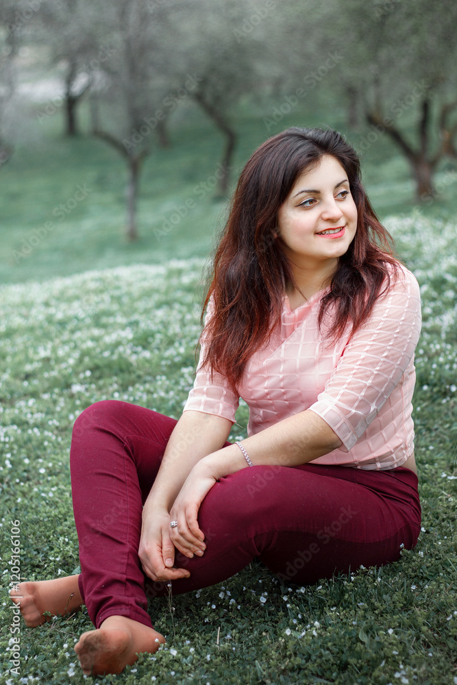 A woman sits barefoot, cross-legged, and looks to the side with a smile on her face. She has long dark hair. Trees with blurred outlines are visible in the background. A woman sits barefoot, cross-legged, and looks to the side with a smile on her face. She has long dark hair. Trees with blurred outlines are visible in the background.