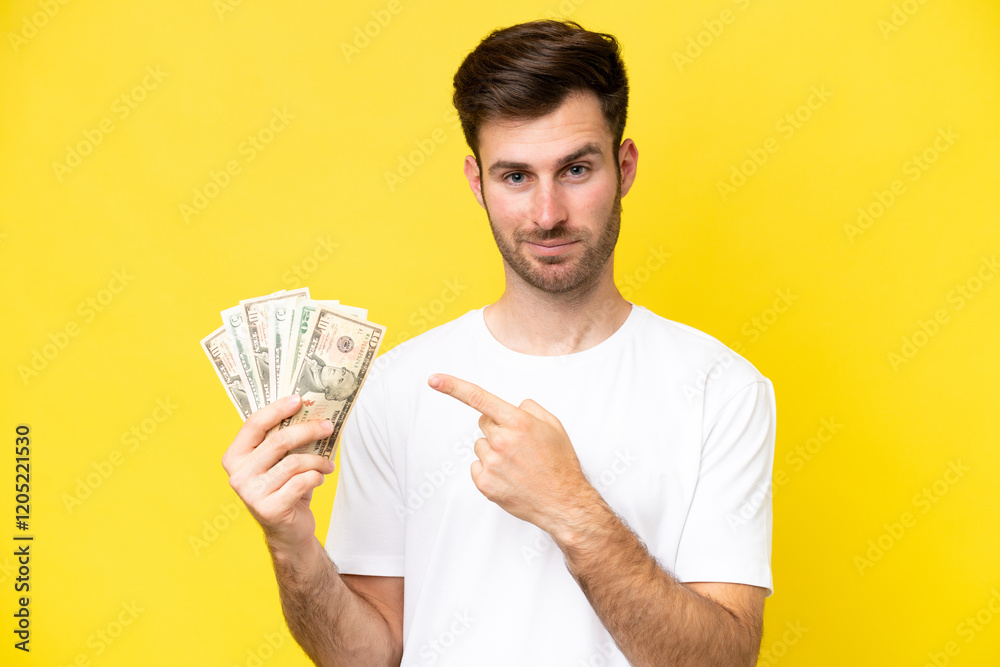 Young caucasian man taking a lot of money isolated on yellow background pointing to the side to present a product Young caucasian man taking a lot of money isolated on yellow background pointing to the side to present a product
