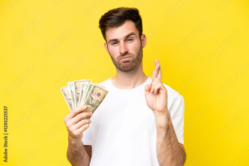 Young caucasian man taking a lot of money isolated on yellow background with fingers crossing and wishing the best Young caucasian man taking a lot of money isolated on yellow background with fingers crossing and wishing the best