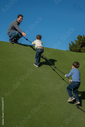 Father helping children climbing rope on artificial turf hill in playground