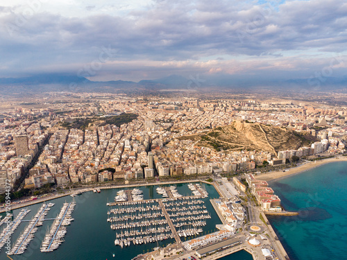 Aerial perspective of Alicante city. Panoramic view of city and port area. Famous travel destination in Spain. Yachts and boats docked. Sunset point with pink reflection on buildings and mountain. 