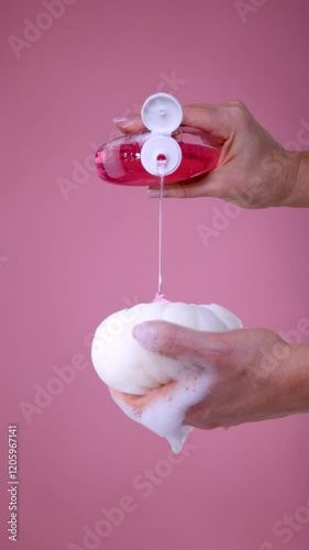 Female hands pouring shower gel from bottle onto white washcloth on pink background