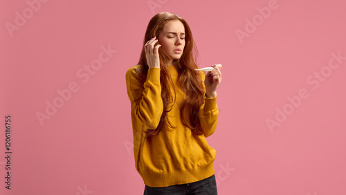 Young woman in her 20s feeling sick, measuring temperature, suffering from fever during seasonal cold on pink studio background. Concept of illness, medicine, treatment, health care