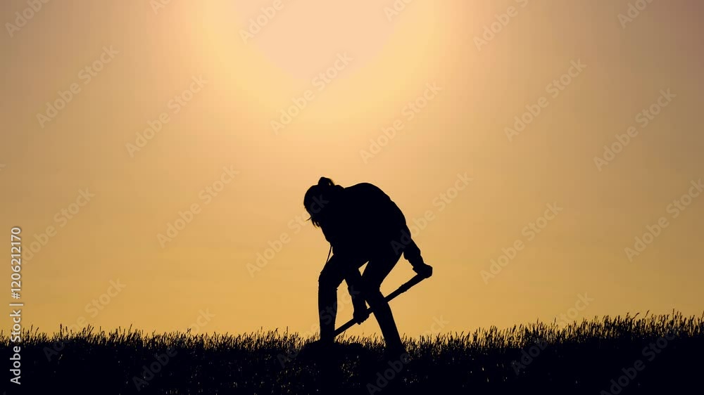Woman digging hole with shovel silhouette in field at sunset light. Woman digs trench in grassy field against sky lit by sunset. Woman farmer works digging soil with shovel in field in evening