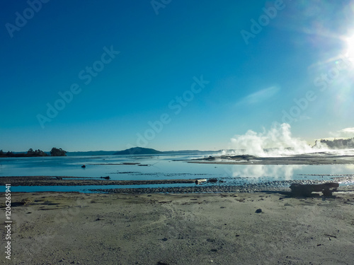 Geothermal area with steam rising from vents and hot springs along shoreline of Rotorua lake, Bay of Plenty Region, North Island New Zealand. Sky is clear blue. mineral-rich wet sand and mudflats