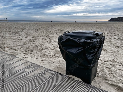 A black trash bin wrapped in a plastic liner stands on a sandy beach near a wooden boardwalk under a cloudy sky. Seaside preservation concept photo