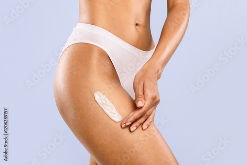 Treatment for Flawless Ideal Skin. Closeup cropped view of slim woman in white underwear panties applying body cream on hip isolated over blue studio background. Lady using moisturize lotion for legs