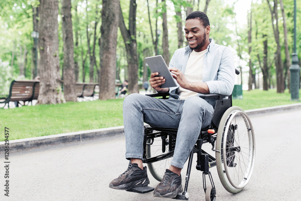 Positive young black guy in wheelchair using tablet computer, browsing web or watching video at urban park, copy space. Full length of African American man checking social media on touch pad outdoors Positive young black guy in wheelchair using tablet computer, browsing web or watching video at urban park, copy space. Full length of African American man checking social media on touch pad outdoors