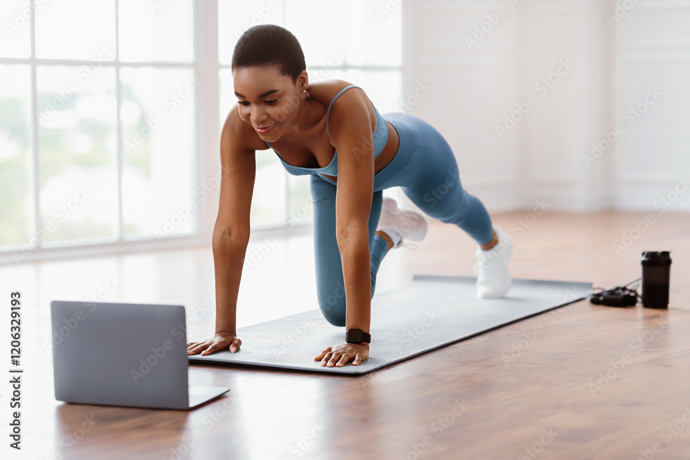 Fototapeta premium Online Training Concept. Sporty African American lady doing mountain climbers exercise, watching video tutorial or having online class on computer, looking at screen, exercising in spatious apartment