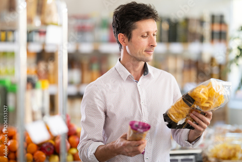 Man in light shirt took some varieties of pasta from shelf. Client compare pasta noodles items package. Customer examines contents of package, choose noodles garnish product photo