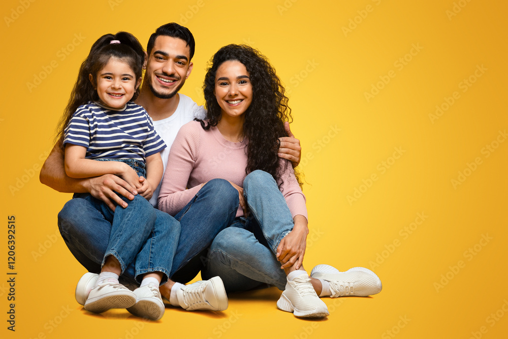 Happy middle eastern family sitting on floor and embracing, cheerful loving arab parents posing together with their cute little daughter over yellow studio background, looking at camera, copy space Happy middle eastern family sitting on floor and embracing, cheerful loving arab parents posing together with their cute little daughter over yellow studio background, looking at camera, copy space