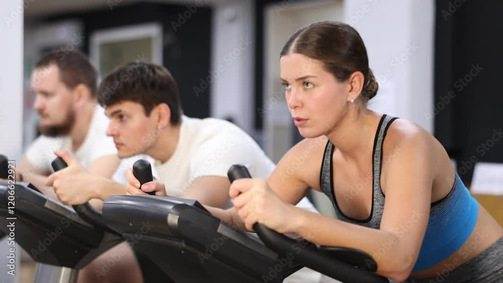 In gym, girl does exercises using stationary bicycle. Young group of athletes trains in gym. Physical education and sports, active healthy lifestyle. In gym, girl does exercises using stationary bicycle. Young group of athletes trains in gym. Physical education and sports, active healthy lifestyle.