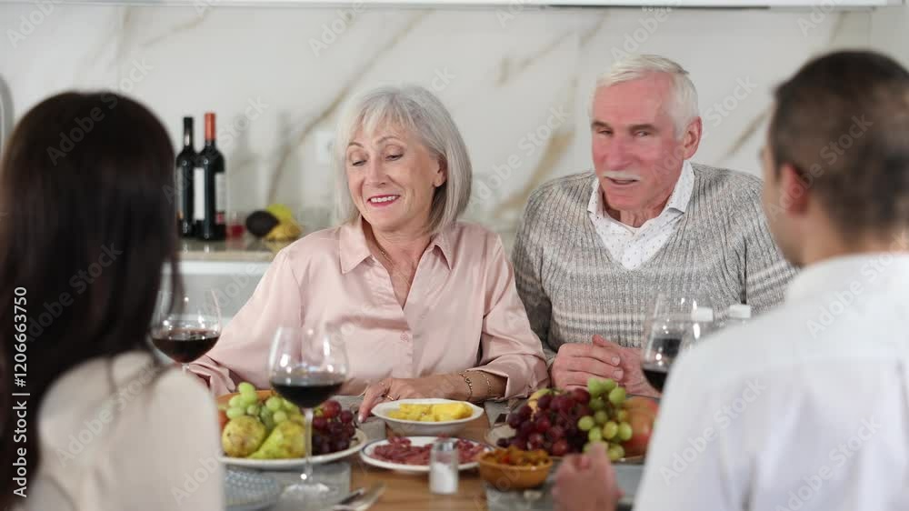 Mature spouses sitting at table, enjoying food and wine, chatting talking laughing with adult son and his girlfriend. In homely atmosphere male and female pensioner celebrate anniversary with guests Mature spouses sitting at table, enjoying food and wine, chatting talking laughing with adult son and his girlfriend. In homely atmosphere male and female pensioner celebrate anniversary with guests