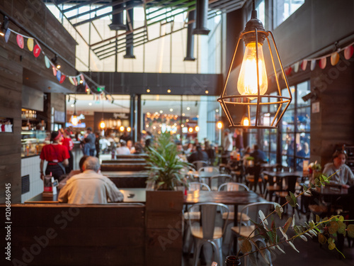 Close of an industrial-style pendant lighting bulb against a blurred view of a bustling casual restaurant with diners enjoying the communal atmosphere and a warm ambiance.