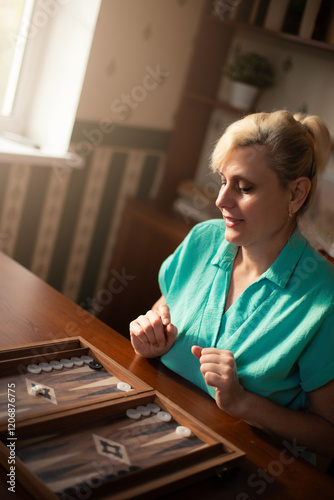 A woman in a turquoise shirt is playing backgammon. She is smiling and focused on the game, with dice and game pieces visible on the wooden board. Natural light fills the warm, cozy setting. photo