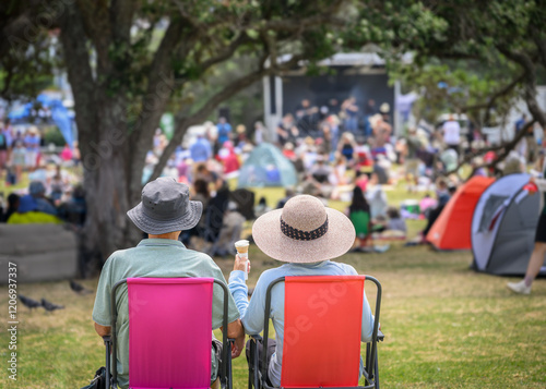 Couple sitting on camping chairs, unrecognizable crowds listen to the music concert in the park. Auckland. photo