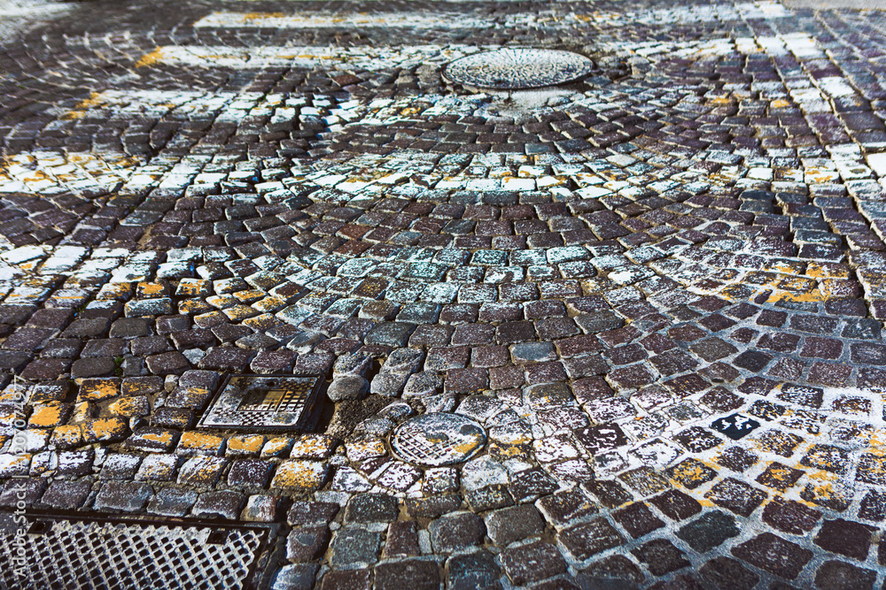Cobblestone street with a circular pattern, manhole covers and drainage grates. Cobblestones are marked with white and yellow paint indicating traffic or pedestrian guidelines