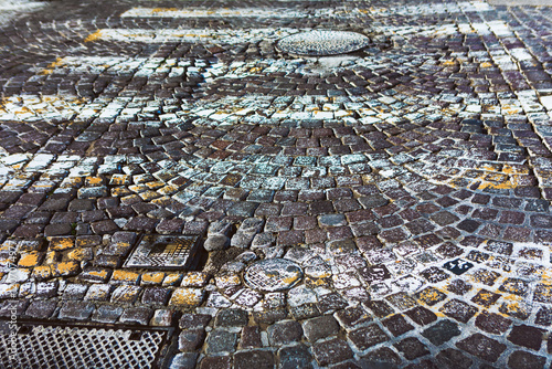 Obraz na płótnie Cobblestone street with a circular pattern, manhole covers and drainage grates. Cobblestones are marked with white and yellow paint indicating traffic or pedestrian guidelines w salonie