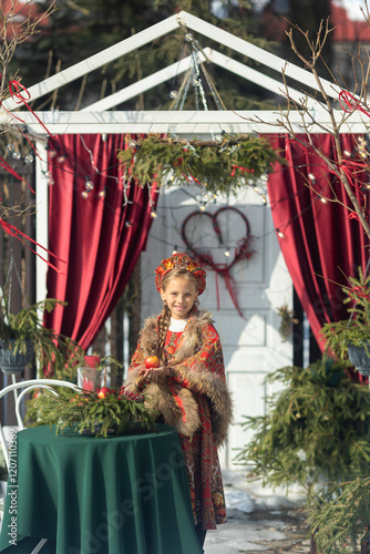 A blonde girl in a national Russian costume on the Maslenitsa holiday. A child in a traditional costume with fur trim and a headdress against a festive backdrop. Winter scene with a beautiful girl