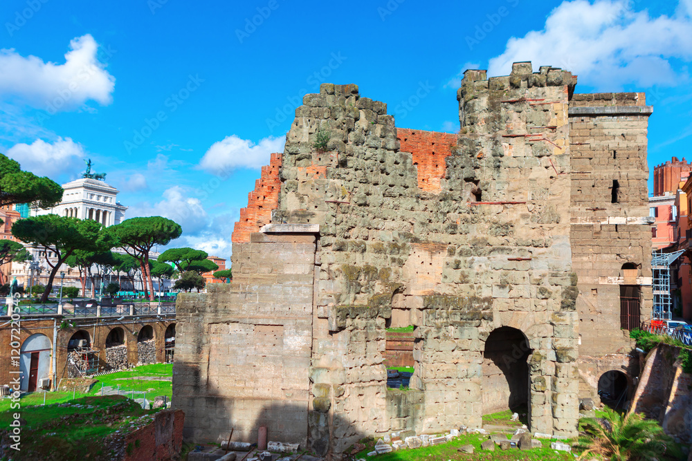 Ancient ruins in Rome, Italy, featuring weathered stone walls under a vivid blue sky, surrounded by lush greenery and iconic umbrella pines. The Altare della Patria is visible in the background